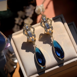 Può includere: Un paio di orecchini pendenti color oro. La parte superiore presenta un design decorativo con cristalli trasparenti. Ogni orecchino ha un grande pendente di cristallo blu a forma di goccia. Gli orecchini sono esposti su una superficie beige.