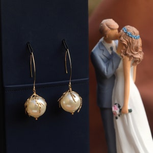 Può includere: Orecchini pendenti color oro con grandi perle finte bianche crema. Le perle sono incastonate in un delicato design a forma di ramo. Gli orecchini pendono da sottili fili curvi. Un cake topper nuziale di una coppia che si bacia è sullo sfondo.