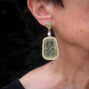 Peut inclure: Boucles d'oreilles dorées avec une pierre centrale verte rectangulaire ornée d'un motif élaboré. Les boucles d'oreilles comportent une petite pierre verte en haut, une perle et sont bordées de petits cristaux transparents. Bijou.