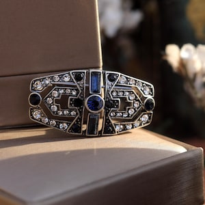 Può includere: Un fermaglio argentato con un design geometrico con strass neri e blu. Il fermaglio ha una pietra blu centrale ed è circondato da strass bianchi più piccoli.