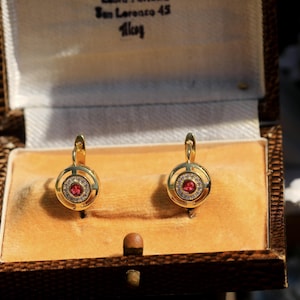 May include: A pair of gold-tone earrings with a circular design. Each earring features a central red gemstone surrounded by smaller clear stones. The earrings are displayed in a vintage jewelry box with a tan interior.