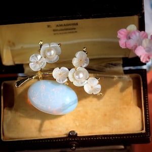 May include: A vintage brooch with a floral design, featuring white petals and pearl accents. The brooch is set on a gold-toned stem, with a light blue oval stone at the base. The brooch is displayed inside a vintage jewelry box.