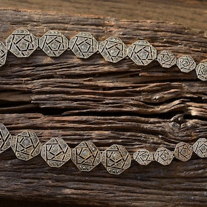 Op de afbeelding: Een gouden ketting met een geometrisch ontwerp. De ketting is gemaakt van meerdere verbonden achthoekige vormen, elk met een reliëf, getextureerd oppervlak en een centraal ruitvormig ontwerp. De ketting ligt op een houten oppervlak.