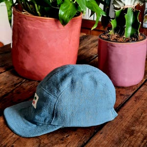 May include: A light blue cap with a small square patch featuring a red and white design. The cap is made of a textured fabric and has a curved brim. The cap is resting on a wooden surface, with two potted plants in the background.
