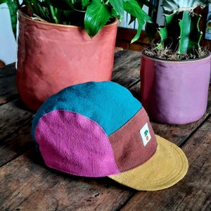 Peut inclure: Casquette à cinq panneaux multicolore avec un dessus turquoise, un panneau latéral magenta et un panneau avant marron. La visière est jaune moutarde et un petit écusson avec un motif vert et orange est à l'avant. La casquette est posée sur une surface en bois.