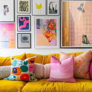 May include: A vibrant living room scene featuring a yellow velvet sofa adorned with colorful throw pillows. Artwork in black frames hangs on the white wall above. The art includes abstract prints and a painting of a shower.