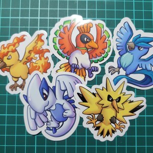 May include: Five colorful cartoon Pokemon stickers featuring the characters: Moltres, Articuno, Zapdos, Flareon, and Jolteon. The stickers are on a green grid background.