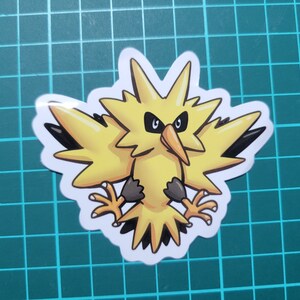 May include: A yellow and black cartoon sticker of a bird-like Pokemon character with a lightning bolt design. The character has a fierce expression with black eyes and a sharp beak.