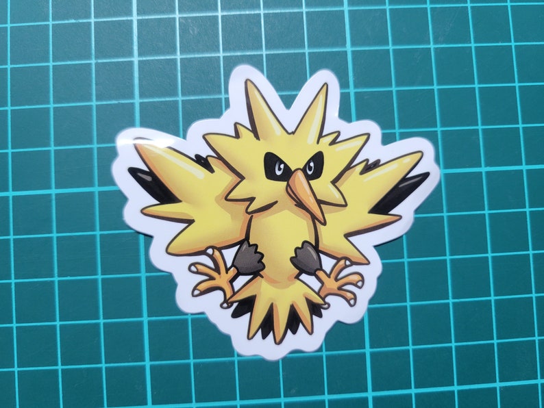 May include: A yellow and black cartoon sticker of a bird-like Pokemon character with a lightning bolt design. The character has a fierce expression with black eyes and a sharp beak.