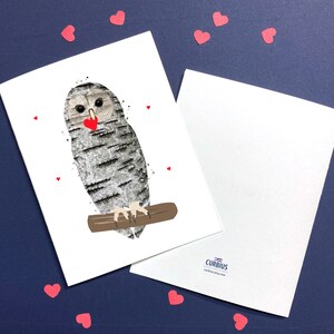 May include: Greeting card featuring an owl with a birch bark design, holding a red heart. The owl is perched on a brown branch, with small red heart accents. A second blank card is visible. The background is a dark blue.