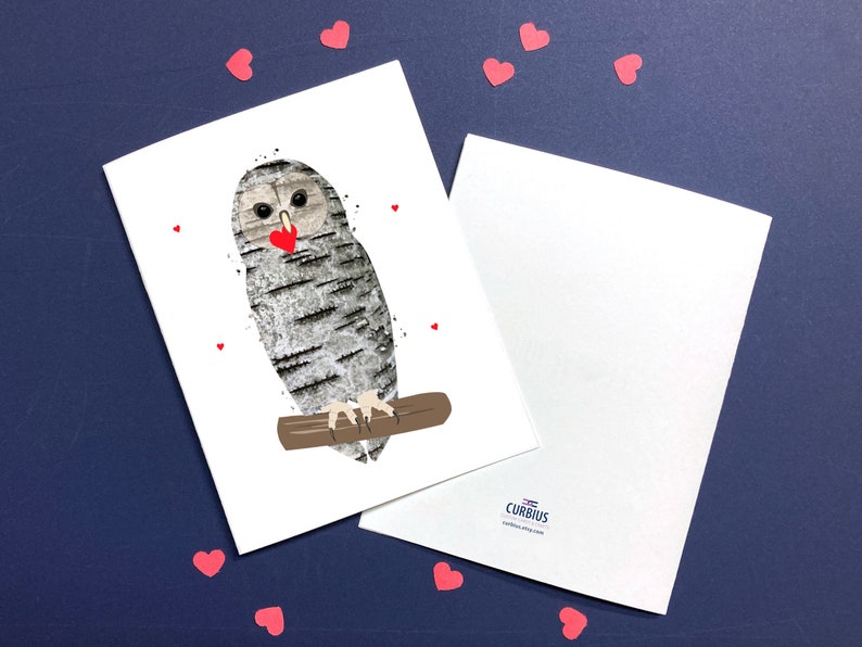 May include: Greeting card featuring an owl with a birch bark design, holding a red heart. The owl is perched on a brown branch, with small red heart accents. A second blank card is visible. The background is a dark blue.