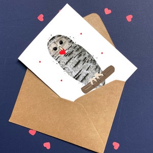 May include: A greeting card featuring an owl illustration with a heart-shaped beak, perched on a branch. The card is inside a brown envelope, with small red heart-shaped confetti scattered around on a dark blue surface.