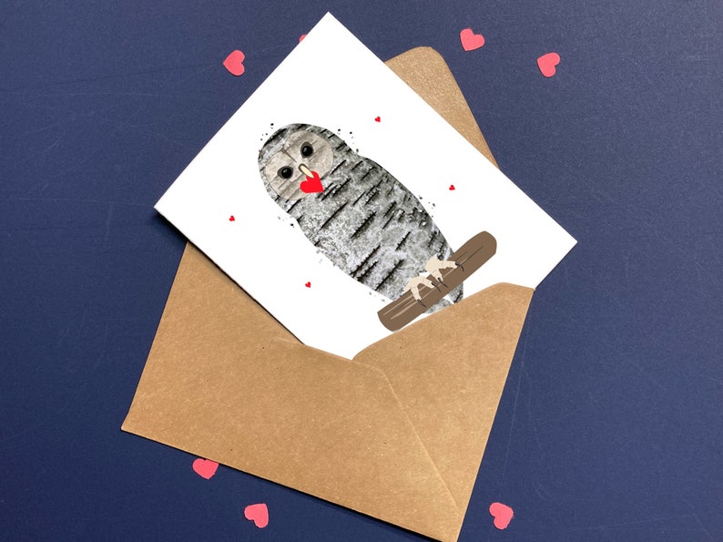 May include: A greeting card featuring an owl illustration with a heart-shaped beak, perched on a branch. The card is inside a brown envelope, with small red heart-shaped confetti scattered around on a dark blue surface.