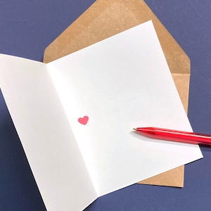 May include: A white greeting card with a small red heart inside a brown envelope, with a red pen resting on the card. The card is open, revealing the blank interior. The background is a dark blue surface.
