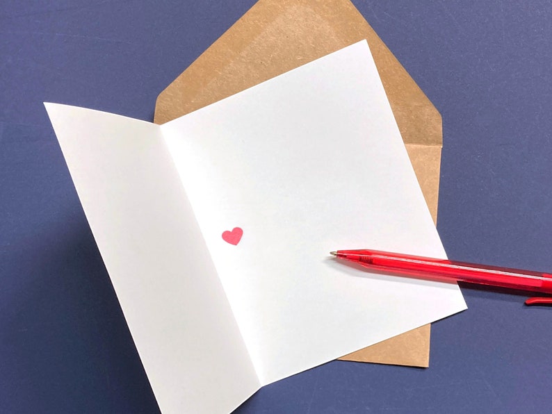 May include: A white greeting card with a small red heart inside a brown envelope, with a red pen resting on the card. The card is open, revealing the blank interior. The background is a dark blue surface.