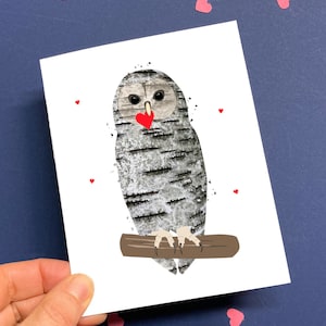 May include: A greeting card featuring an owl illustration. The owl has a birch bark texture and is perched on a brown branch. It holds a red heart in its beak, with small red hearts scattered around it. The card is held against a dark blue background.