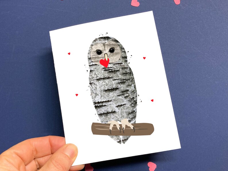 May include: A greeting card featuring an owl illustration. The owl has a birch bark texture and is perched on a brown branch. It holds a red heart in its beak, with small red hearts scattered around it. The card is held against a dark blue background.