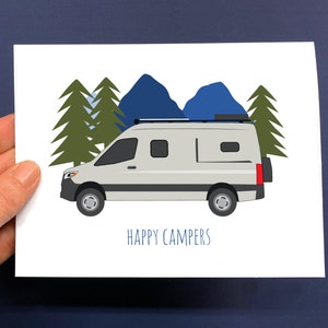 May include: A white card with a drawing of a gray camper van, green trees, and blue mountains. The text "HAPPY CAMPERS" is written in blue below the van. The card is held by a hand.