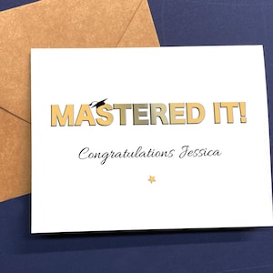 May include: A white graduation card with the words "MASTERED IT!" in gold and gray lettering, topped with a graduation cap illustration. The card also includes the text "Congratulations Jessica" and a small gold star. An open brown envelope is in the background.