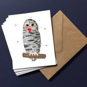 May include: A stack of white greeting cards featuring an owl illustration. The owl has a birch bark pattern, a red heart, and is perched on a brown branch. A brown envelope is next to the cards on a dark blue surface.