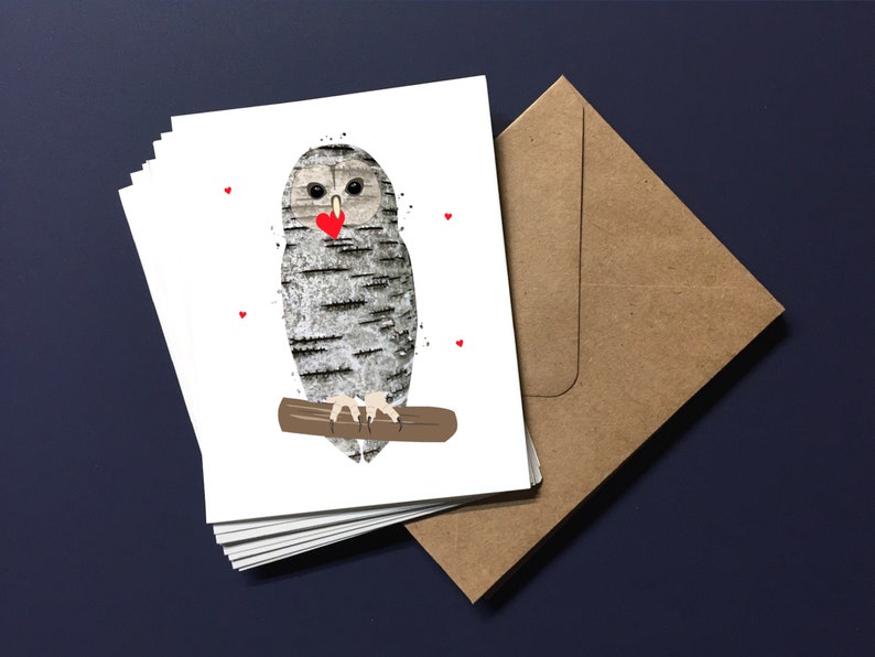 May include: A stack of white greeting cards featuring an owl illustration. The owl has a birch bark pattern, a red heart, and is perched on a brown branch. A brown envelope is next to the cards on a dark blue surface.