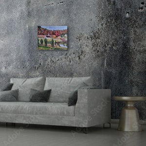 May include: A painting of a small town with a church in the center, surrounded by trees with fall foliage. The painting is framed and hanging on a gray concrete wall above a gray couch.