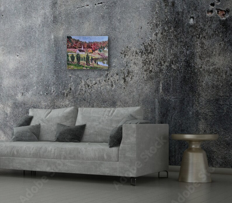 May include: A painting of a small town with a church in the center, surrounded by trees with fall foliage. The painting is framed and hanging on a gray concrete wall above a gray couch.