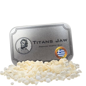May include: A silver tin with the words "Titans Jaw Premium Mastic Gum" printed on it. The tin is open and a pile of white, opaque, irregularly shaped mastic gum pieces are spilling out.