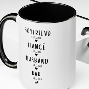 K&ouml;nnte beinhalten: Eine wei&szlig;e Keramiktasse mit schwarzem Henkel und schwarzem Rand. Die Tasse hat schwarzen Text, der "BOYFRIEND EST. 2014", "FIANC&Eacute; EST. 2019", "HUSBAND EST. 2020" und "DAD EST. 2023" lautet.