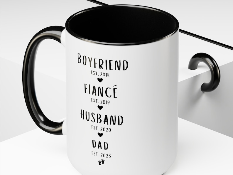 K&ouml;nnte beinhalten: Eine wei&szlig;e Keramiktasse mit schwarzem Henkel und schwarzem Rand. Die Tasse hat schwarzen Text, der "BOYFRIEND EST. 2014", "FIANC&Eacute; EST. 2019", "HUSBAND EST. 2020" und "DAD EST. 2023" lautet.