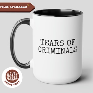 May include: White ceramic mug with a black rim and handle. The mug has the text "TEARS OF CRIMINALS" printed in black on the front. The mug is sitting on a gray surface.