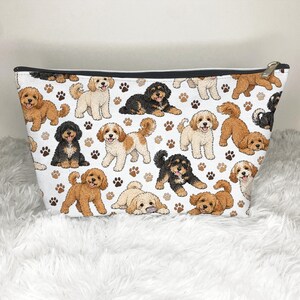May include: A white zipped pouch featuring a repeating pattern of illustrated doodle dogs in shades of cream, tan, brown, and black, interspersed with small brown paw prints. The bag has a black top trim and a silver-toned metal zip pull.