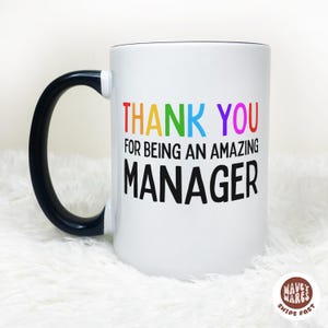 Op de afbeelding: Witte keramische mok met een zwarte handgreep en rand. De mok heeft de tekst "THANK YOU FOR BEING AN AMAZING MANAGER" in kleurrijke letters. Een cadeau voor een manager.