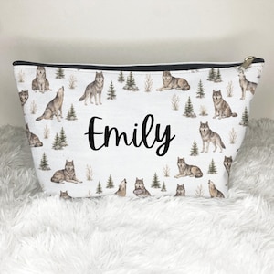 May include: A white zippered pouch featuring a repeating pattern of gray wolves and green pine trees. The name Emily is printed in large black script in the center of the pouch. The bag has a black zipper closure at the top edge.