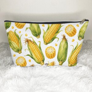 May include: A zippered pouch featuring a repeating pattern of yellow corn cobs with green husks and individual corn kernels on a white background. The pouch has a black top edge and a silver-toned metal zipper pull at the top right corner.