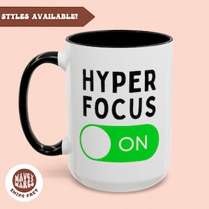 Op de afbeelding: Een witte keramische koffiemok met een zwarte handgreep. De mok heeft zwarte tekst die "HYPER FOCUS" luidt met een groene schakelaar die "ON" zegt.