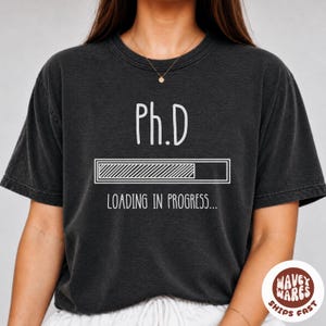 以下が含まれることがあります： チャコールグレーの半袖Tシャツ。中央には白い文字で「Ph.D」と書かれ、その下に斜線で部分的に塗りつぶされた読み込みバーのグラフィックがあり、さらにその下に「LOADING IN PROGRESS...」という白い文字が配置されています。
