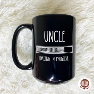 May include: Black ceramic mug with a large handle. The mug features the word "UNCLE" in white, above a loading bar graphic, and the text "LOADING IN PROGRESS..." below. The mug has a glossy finish.