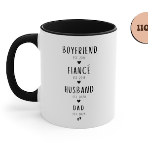 K&ouml;nnte beinhalten: Eine wei&szlig;e Keramiktasse mit schwarzem Rand und Henkel. Die Tasse hat einen schwarzen Text mit der Aufschrift "Boyfriend Est. 2014", "Fianc&eacute; Est. 2019", "Husband Est. 2020" und "Dad Est. 2025".