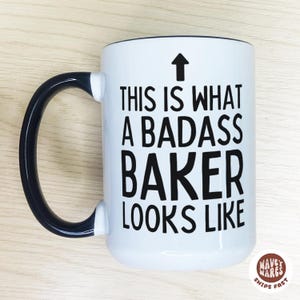 May include: White ceramic mug with a black handle and rim. The mug features the text "THIS IS WHAT A BADASS BAKER LOOKS LIKE" in black, with an upward-pointing arrow above the text. The mug has a glossy finish.