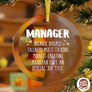 Puede incluir: Adorno navideño de cristal transparente con cinta dorada. El adorno tiene la palabra "MANAGER" en blanco, junto con el texto "BECAUSE BADASS TALENTED MULTI-TASKING MIRACLE-CREATING MAGICIAN ISN'T AN OFFICIAL JOB TITLE."