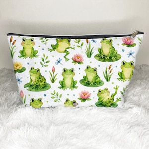 May include: A white zippered pouch featuring a repeating watercolor pattern of green frogs, pink lotus flowers, green lily pads, cattails, and blue dragonflies. The bag has a black zipper closure along the top edge and a silver-toned metal zipper pull.