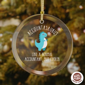 May include: A clear, round Christmas ornament with a gold ribbon for hanging. The ornament features the text "Accountasaurus" above a cartoon dinosaur and the phrase "Like a normal accountant, but cooler."