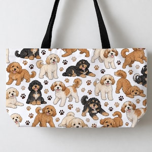 Puede incluir: Un bolso tote blanco con correas negras para el hombro que presenta un patrón repetitivo de perros doodle ilustrados en varios colores, incluyendo crema, marrón dorado y negro, intercalados con pequeños iconos de huellas en marrón y negro.