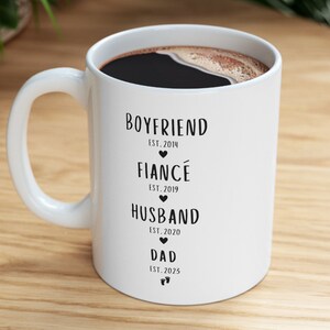 K&ouml;nnte beinhalten: Wei&szlig;e Keramiktasse mit schwarzem Text, der "Boyfriend Est. 2014", "Fianc&eacute; Est. 2019", "Husband Est. 2020" und "Dad Est. 2025" lautet.