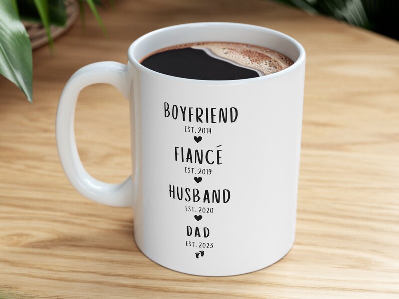 K&ouml;nnte beinhalten: Wei&szlig;e Keramiktasse mit schwarzem Text, der "Boyfriend Est. 2014", "Fianc&eacute; Est. 2019", "Husband Est. 2020" und "Dad Est. 2025" lautet.