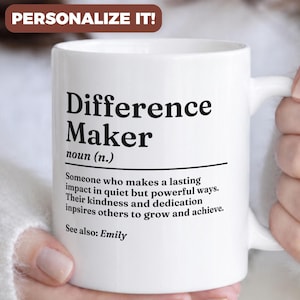 Può includere: Tazza in ceramica bianca con la scritta "Difference Maker" in nero, insieme a una definizione. La tazza include anche la scritta "See also: Emily". La tazza ha un manico ed è tenuta da una persona.