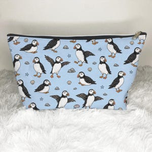 May include: A light blue zippered pouch featuring a repeating pattern of black and white puffins with orange beaks and feet. The design includes small illustrations of gray rocks, tan seashells, and blue starfish scattered among the puffins.
