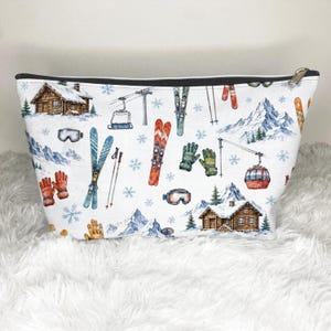 May include: White cosmetic bag with a black zipper and a winter ski theme. The bag features watercolor illustrations of skis, ski lifts, snow-covered cabins, mountains, snowflakes, and ski goggles.