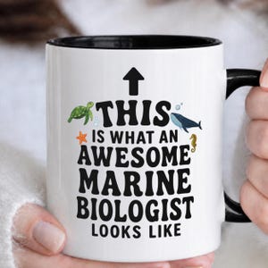 Puede incluir: Taza de cerámica blanca con interior y asa negros. La taza presenta el texto "THIS IS WHAT AN AWESOME MARINE BIOLOGIST LOOKS LIKE" con ilustraciones de una tortuga, una ballena, una estrella de mar y un caballito de mar.